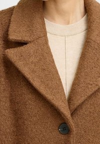 Bruine textuurjas met een gerafelde kraag en een zwarte knoop, gedragen over een licht beige gebreide top. Close-up weergave.