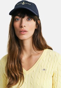 Marineblaue Baseballcap mit goldenem Ankerlogo; das Modell trägt einen hellgelben, grob gehäkelten Pulli mit tiefem V-Ausschnitt und kleinem Logo auf der linken Brust.