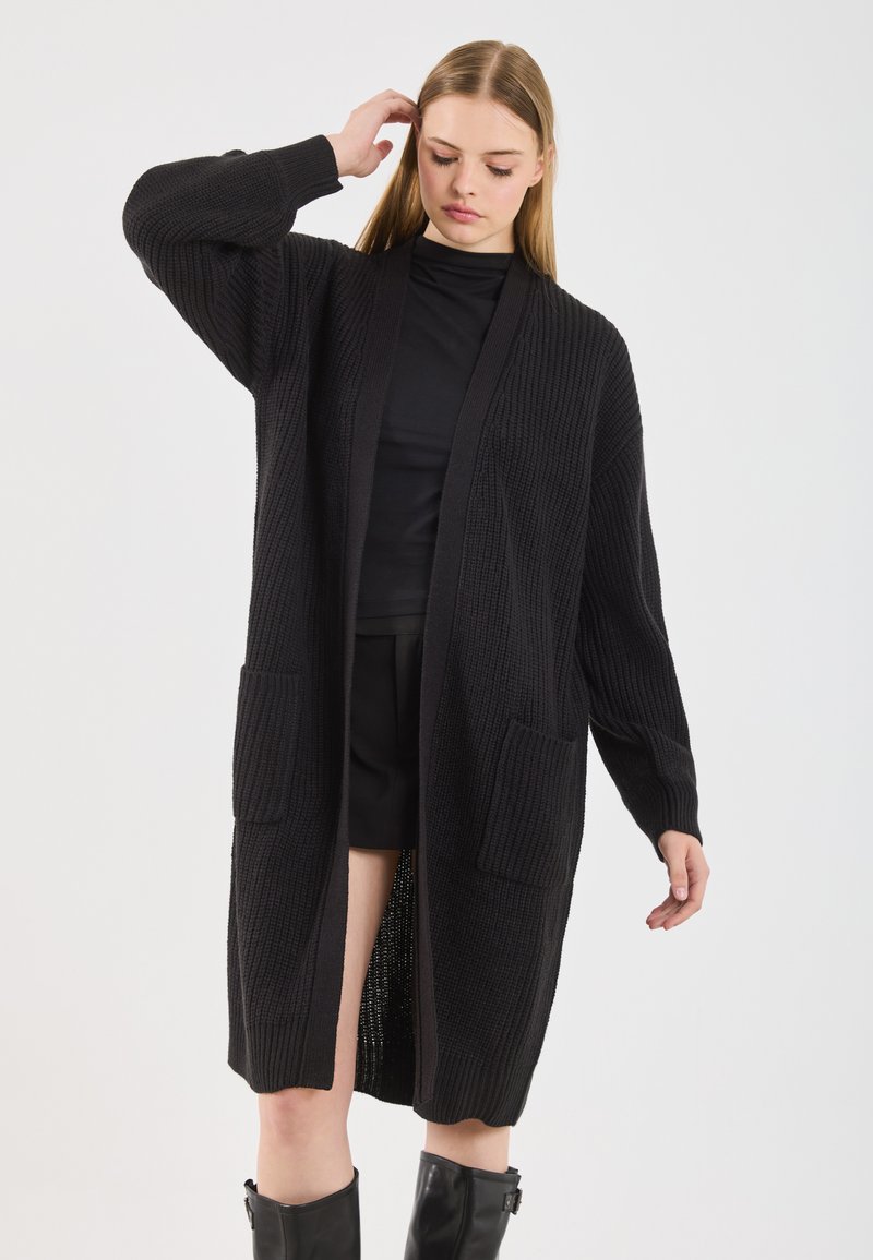 Jovem mulher vestindo um longo cardigan de malha preto com bolsos, top preto, calções e botas negras até ao joelho, contra um fundo branco.
