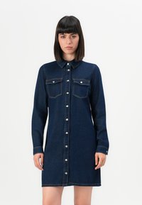 Jeune femme portant une robe en denim bleu foncé à boutons avec des poches avant, se tenant devant un fond blanc uni.