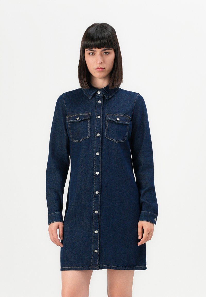 Jeune femme portant une robe en denim bleu foncé à boutons avec des poches avant, se tenant devant un fond blanc uni.