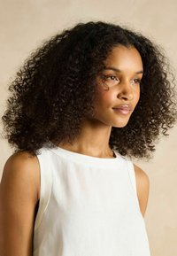 Model mit dunklem, lockigem Haar, die ein ärmelloses weißes Oberteil trägt. Sanfte Textur, natürlicher Look, vor einem neutralen Hintergrund.