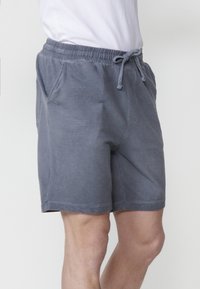 Man som bär grå texturerade shorts med snörning och en vit skjorta, stående mot en enkel ljus bakgrund.