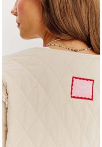 Beżowa kamizelka pikowana z łatką w czerwonej ramce z logo "NAOKO" i falbaniastymi brzegami. Naszyjnik z koralików dodaje kolorowych akcentów.