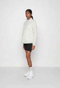 Svetlosivá mikina na zips s jemnou textúrou, nosená na čiernych krátkych nohaviciach, s vysokým golierom a voľným strihom. Celý outfit dopĺňajú biele tenisky.