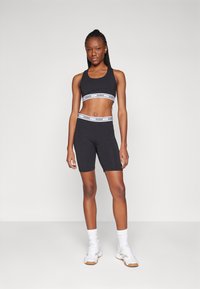 Ensemble de sport noir comprenant un top court et un short de cyclisme, tous deux ornés de bandes blanches avec le logo "GUESS", fabriqué à partir d'un tissu lisse et extensible.