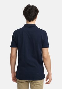 Marineblå polo skjorte med korte ermer og krage, laget av mykt stoff. Har en ren, klassisk skjæring med en rett kant bak.