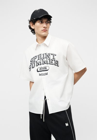 Jeune homme portant une chemise blanche à manches courtes avec le texte "SPRINT SUMMER 2026 MSGM", un pantalon noir à bordure blanche et une casquette noire, regardant sur le côté.