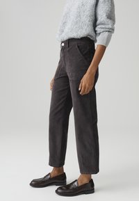 Personne portant un pull gris duveteux, un pantalon gris foncé taille haute et des mocassins en cuir brun foncé, debout sur un fond uni.