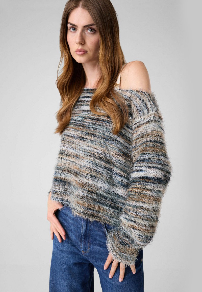 Jeune femme aux longs cheveux portant un pull duveteux multicolore avec une épaule découverte et un jean bleu, debout devant un fond uni.