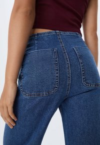 Žena v modrých džínách s vysokým pasem a velkými zadními kapsami, oblečená v bordó krátkém topu, na jednolitém pozadí.