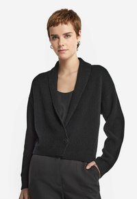 Svart ribbad cardigan med sjalkrage, enkel knappstängning och sänkta axlar. Bärs över en enkel svart topp, visad mot en neutral bakgrund.