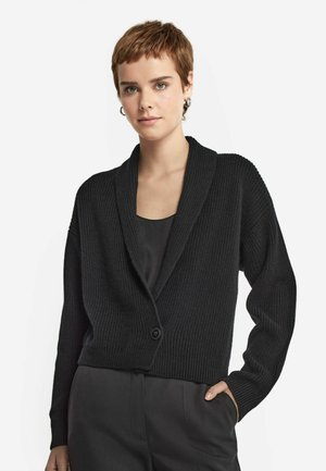 Sort ribbet cardigan med sjalskrave, enkelt knaplukning og dropped skuldre. Båret over en simpel sort top, vist på en neutral baggrund.