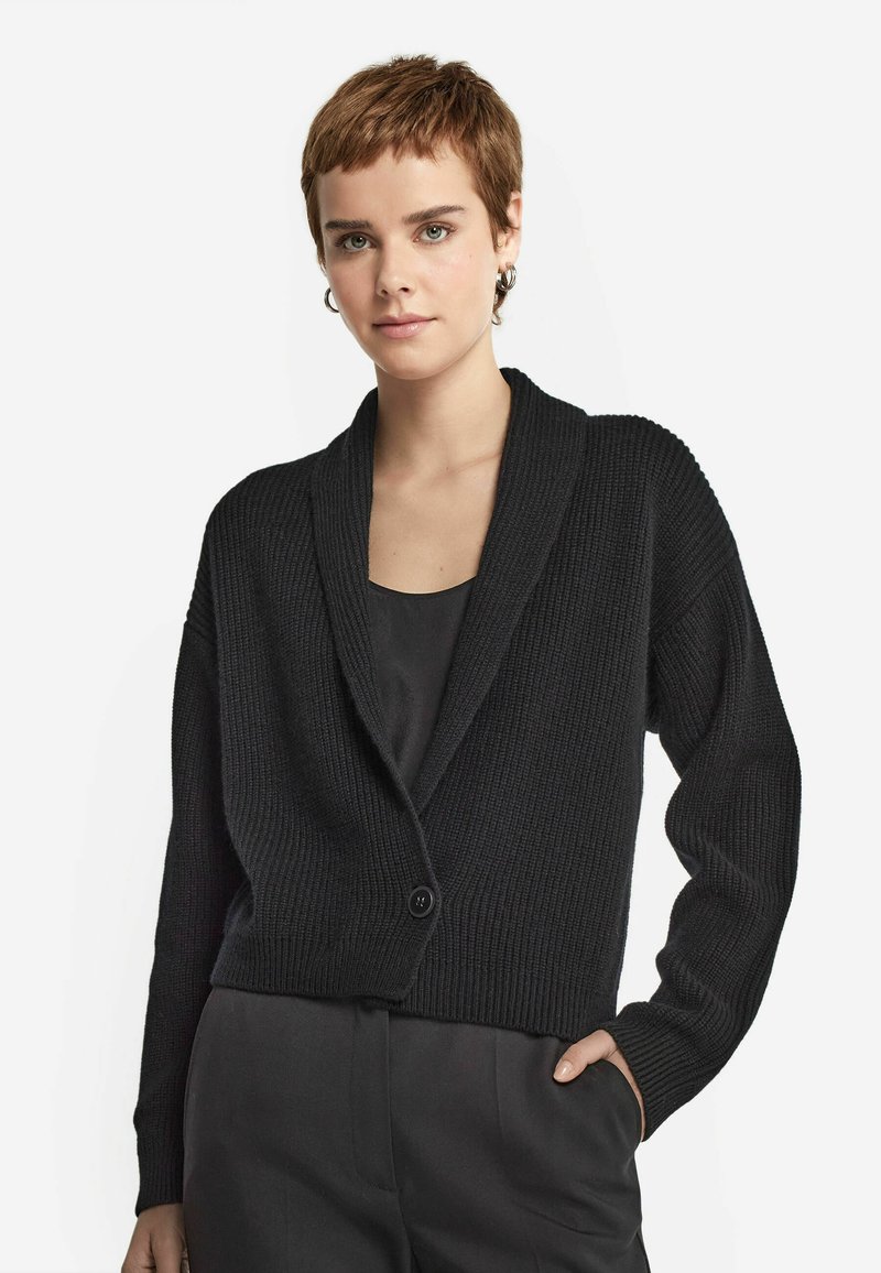 Svart ribbad cardigan med sjalkrage, enkel knappstängning och sänkta axlar. Bärs över en enkel svart topp, visad mot en neutral bakgrund.