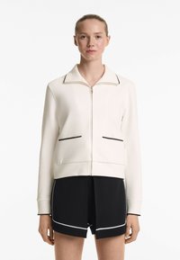 Light beige zip-up jacket with black trim and two front pockets, paired with black shorts featuring white accents. Smooth fabric texture.