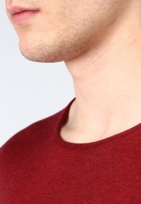 Roter gestrickter Pullover mit rundem Ausschnitt, strukturiertem Stoff und sichtbaren feinen Fasern, getragen von einer Person, die einen Teil ihres Halses und Kinns zeigt.