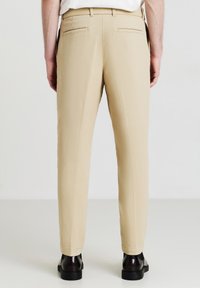 Pantaloni beige sartoriali con vestibilità dritta, caratterizzati da due tasche posteriori e una texture liscia. Indossati con scarpe nere.
