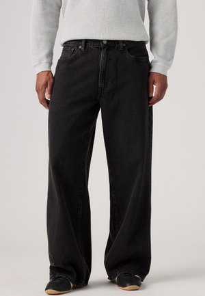 Personne portant un jean noir ample, un t-shirt gris clair à manches longues en maille gaufrée et des baskets noires avec des semelles claires, debout devant un fond uni.