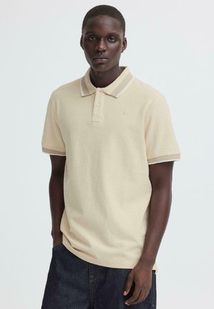 Jeune homme à la peau foncée portant un polo beige à manches courtes et un pantalon en jean foncé, debout devant un fond clair uni.