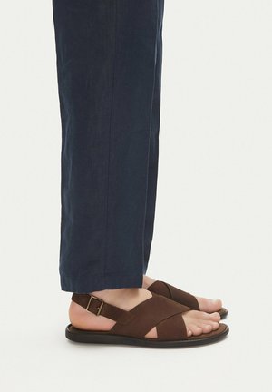 Stopy w brązowych skórzanych sandałach z klamrami, zestawione z granatowymi spodniami, pokazane z boku na białym tle.