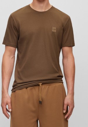 Man wearing brown short-sleeve T-shirt with small logo patch on chest, paired with brown drawstring pants against plain background.
