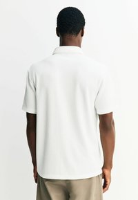 Weißes Poloshirt mit kurzen Ärmeln und strukturiertem Rippmuster, ausgestattet mit einem klassischen Kragen und geradem Saum, kombiniert mit beigen Hosen.