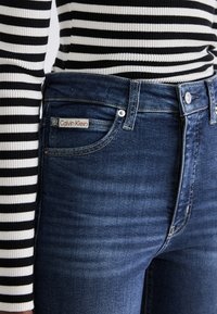 Tumman pesun farkut, joissa on Calvin Klein -logo etutaskussa, yhdistettynä musta-valko raidalliseen pitkähihaiseen paitoihin.