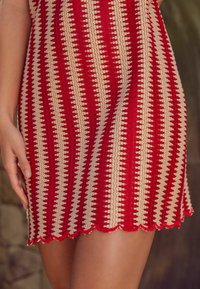 Vestido de ganchillo rojo y beige con un patrón a rayas, dobladillo en picos y de longitud corta. Mano colocada sobre la tela del vestido.