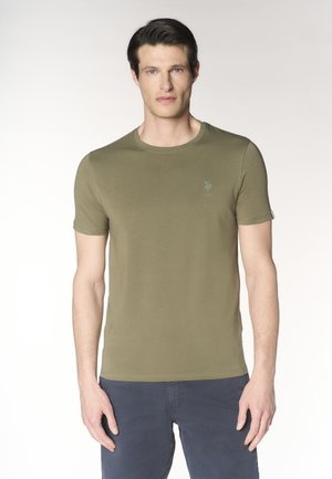 Uomo che indossa una maglietta verde oliva a maniche corte con logo USPA e pantaloni blu scuro, in piedi davanti a uno sfondo chiaro uniforme.