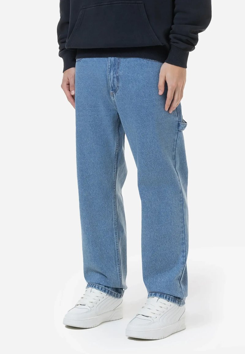 Fatos de ganga azul claro com um ajuste relaxado, apresentando bolsos laterais e um desgaste sutil. Combinados com ténis brancos.