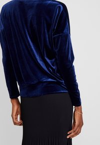 Top in velluto blu navy con maniche lunghe, vestibilità rilassata e texture drappeggiata. Abbinato a una gonna nera plissettata.