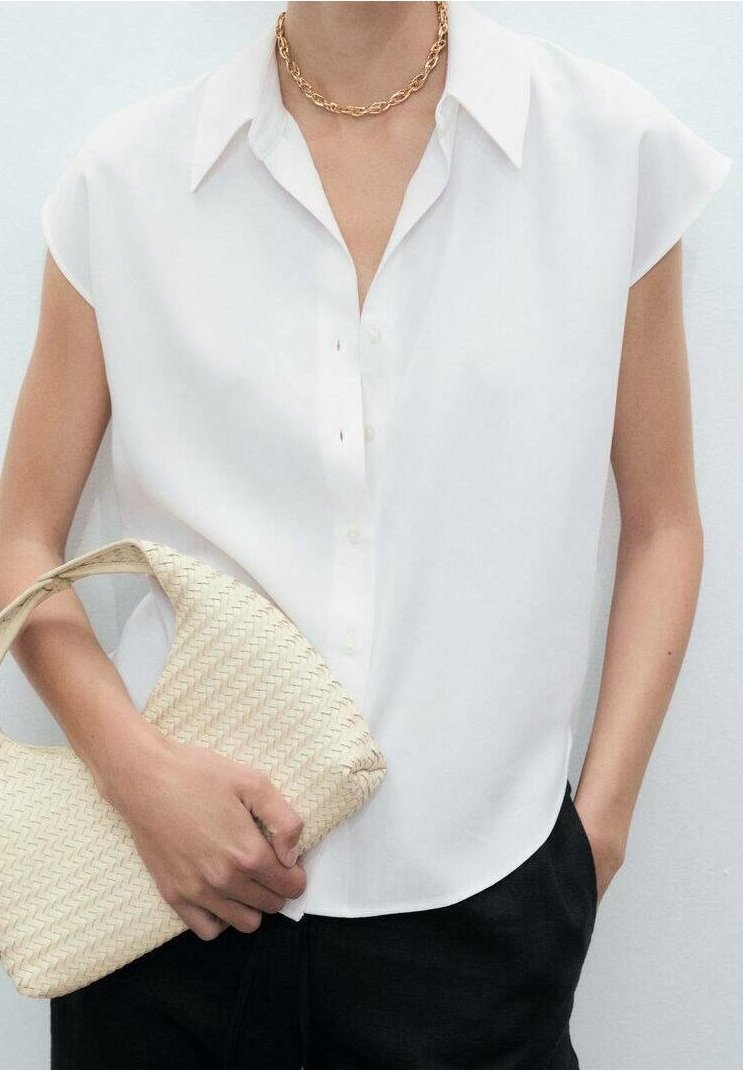 Femme portant un chemisier blanc sans manches et un collier en chaîne dorée, tenant un sac beige tressé, une main dans la poche d'un pantalon noir.