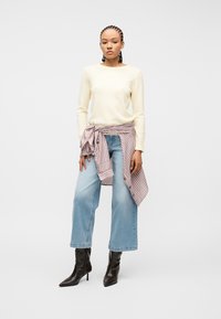 Femme aux cheveux tressés portant un pull crème, une chemise rayée nouée à la taille, un jean à jambes larges et des bottes noires à talons, debout devant un fond blanc.