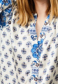 Blusa di colore chiaro con un motivo floreale e geometrico blu. Il tessuto è morbido con scollo a V e dettagli decorativi lungo la parte anteriore.