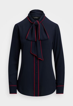 Jūras zilais krekls ar garām piedurknēm, ar tauriņu apkakli, sarkanu apdari pa malām un pogu aizdari ar melnām pogām. Gluda auduma tekstūra.