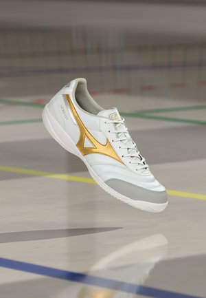 Chaussure de football indoor blanche avec logo doré et bout gris, suspendue au-dessus d'un sol de gym marqué de lignes de terrain.
