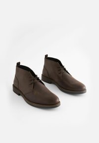 Botas de cuero marrón hasta el tobillo con punta redonda, cordones mínimos y un pequeño tacón. Textura suave con un brillo sutil. Suela de goma plana.