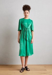 Grünes Satin-Kleid mit kurzen Puffärmeln, gebundenem Taillengürtel und knielangem Saum. Kombiniert mit schwarzen offenen Mules auf einem Holzboden.
