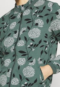 Grön zip-up jacka med blommönster i vitt och svart, texturerad tyg och samlad krage.