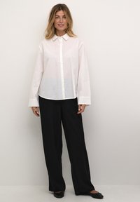 Camisa blanca de botones con mangas largas, que presenta sutiles elementos decorativos. Combinada con pantalones de pierna ancha negros y zapatos planos.