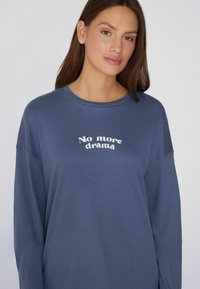 Sudadera azul de manga larga hecha de un tejido suave, con un texto impreso en blanco que dice "No más drama" en un diseño curvado en la parte delantera.