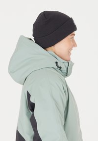 Ljusgrön vattentät jacka med svart huva, med mörkgrå accenter och en ribbad svart mössa som bärs på huvudet av en leende person.