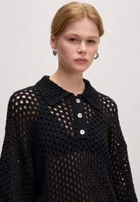 Schwarzes, gehäkeltes Strickoberteil mit einem kragenartigen Ausschnitt, das über eine Knopfleiste und große Ausschnittmuster verfügt. Lange Ärmel mit einem lockeren Schnitt.