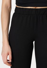 Juoda kelnės su elastinga juosmens juosta, pagamintos iš minkštos medžiagos, pasižyminčios lygiu paviršiumi ir prigludusia forma, su arti matomu žiūrėjimu į juosmens juostą.