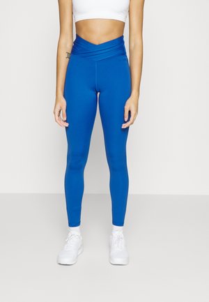 Femme portant des leggings bleus taille haute avec une ceinture croisée, un crop top blanc, des chaussettes blanches et des baskets blanches, debout sur un fond uni.