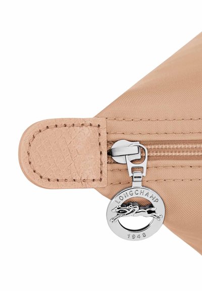 Beige bag corner with beige leather tab, beige zipper, and round silver zipper pull engraved with "LONGCHAMP 1948" and a horse logo.