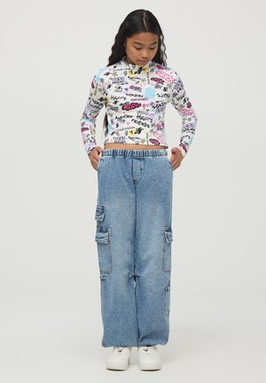 Biała, dopasowana bluzka z długim rękawem z wielokolorowym graficznym napisem; zestawiona z luźnymi, jasnoniebieskimi dżinsami w stylu cargo z kieszeniami. Białe sneakersy.