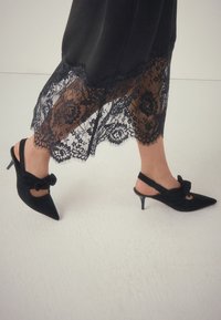 Escarpins noirs en suédine à bride arrière avec bout pointu, ornés d'un nœud et d'un talon stiletto fin, assortis à une jupe bordée de dentelle.