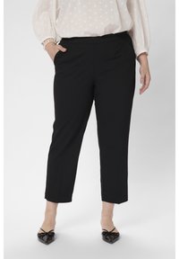 Mujer vistiendo pantalones rectos cortos negros con bolsillos, combinados con zapatos de charol negros de tacón bajo y una blusa blanca texturizada.