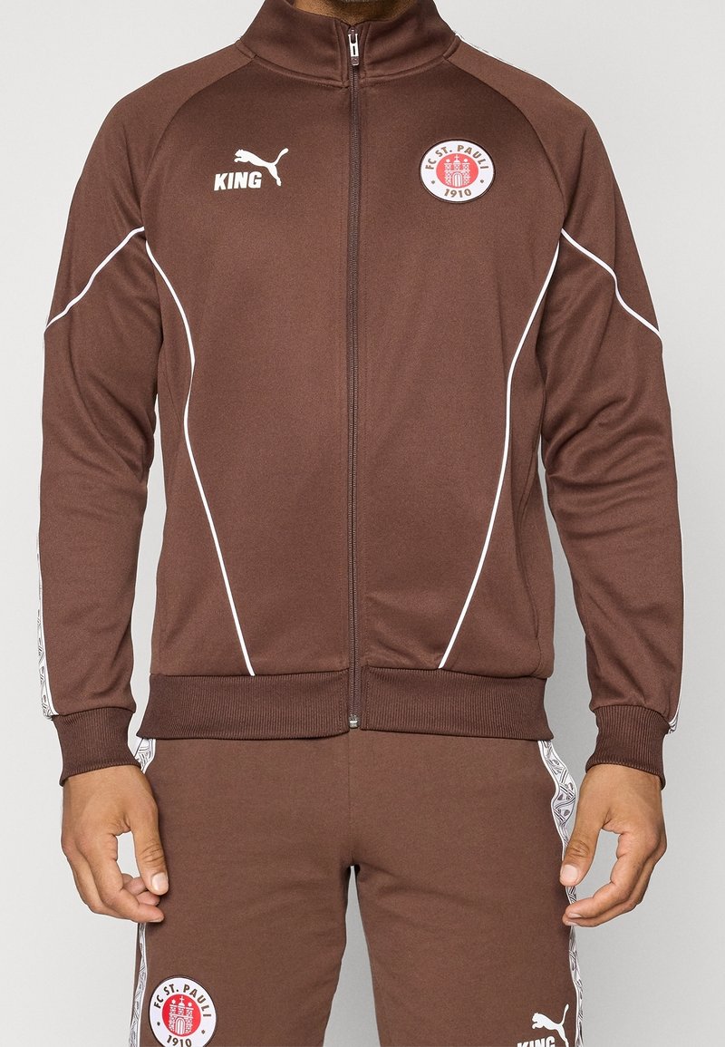 Homme portant une veste et un pantalon de sport marron FC St. Pauli avec des logos blancs Puma et King, debout, bras détendus le long du corps.